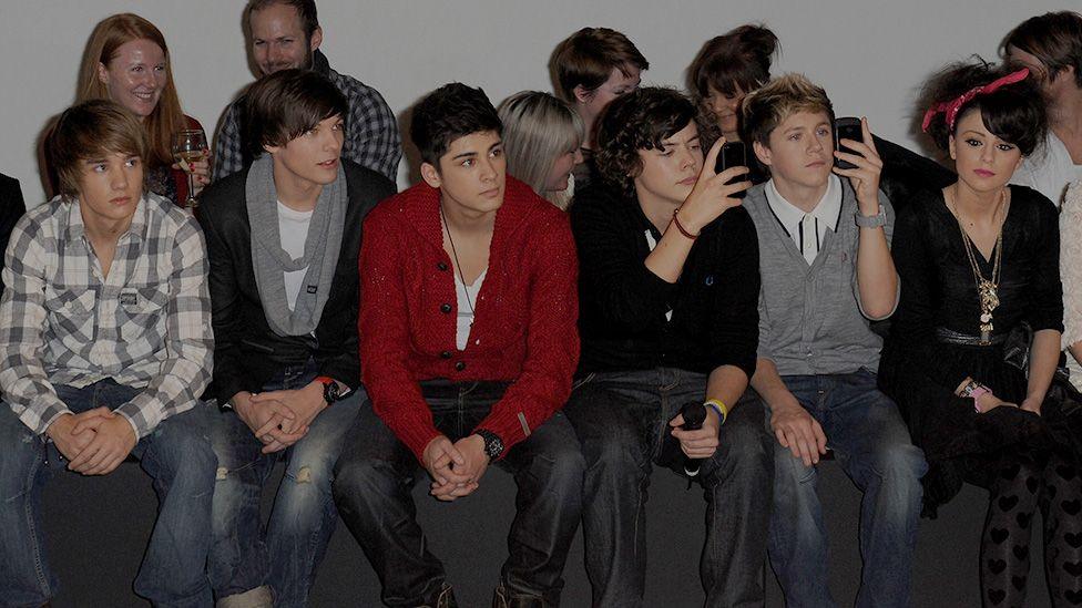 Five young men and a young woman sit in the front row of a catwalk show. The group looks on with interest, with two of the male members holding up their phones to take pictures. The men are dressed in 2010-era fashion - a mixture of checked shirts, chunky cardigans and assymetrical haircuts. The young women uses a red Paisley bandana to tie up her curly black hair, and wears a selection of large gold pendants alongside her all-black dress and top combo. 