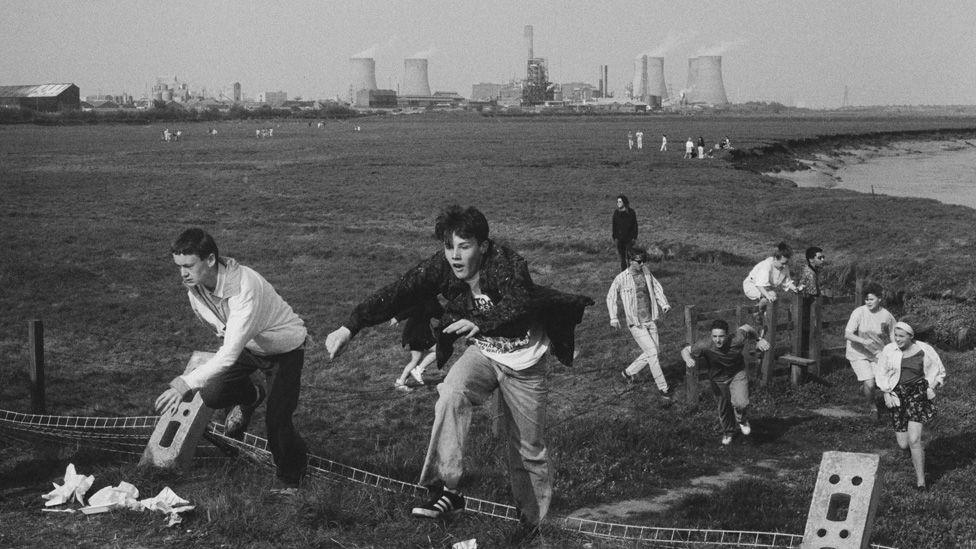 Gatecrashers jump over the remains of the fence, with wateland and an industrial plant in the background