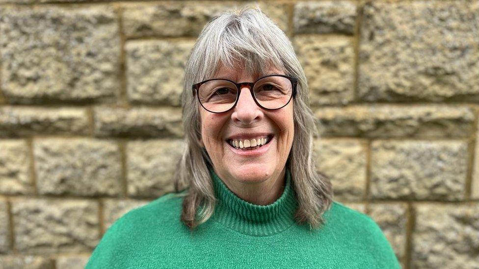 Juliet Layton wearing a green jumper and smiling at the camera in front of a brick wall