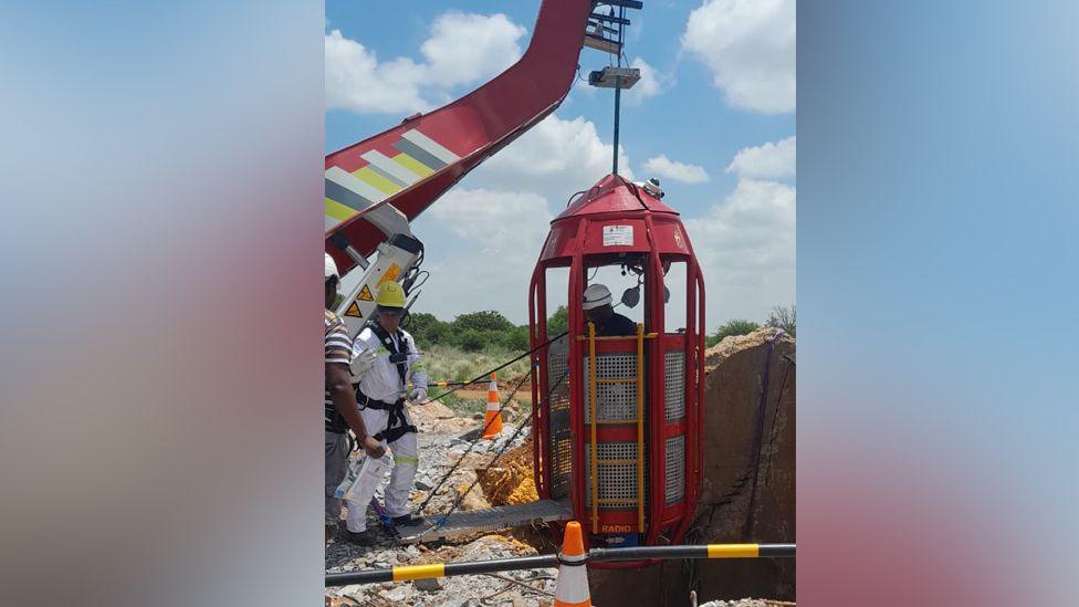 A man in a white boiler stands by a crane that is about to lower a cage down a mine shaft.