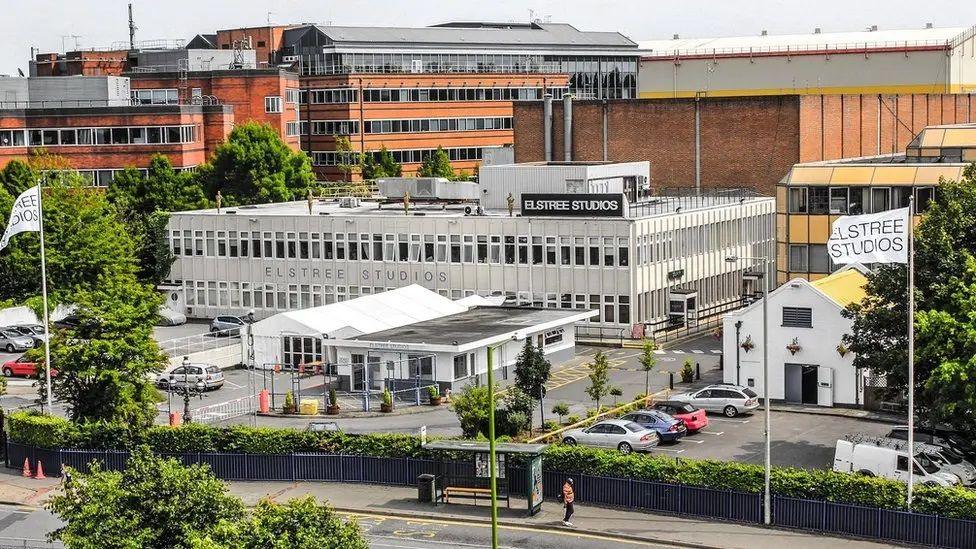 Elstree Studios seen from a distance. It is a white two storey building with a car park at the front and a smaller reception building in the car park. There are two large flagpoles with white flags saying Elstree Studios. Buildings surround it.
