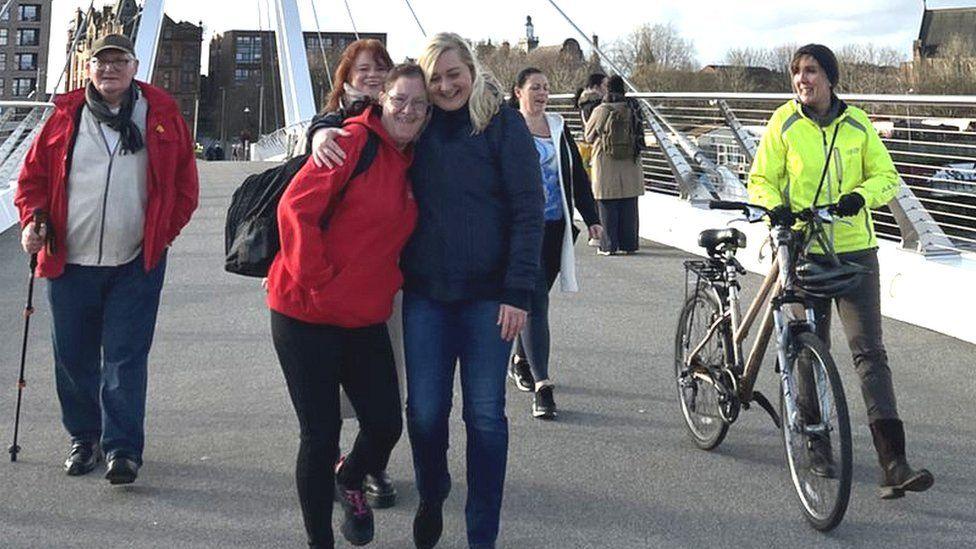 A group of people walking on the new Govan-Partick bridge over the River Clyde. One woman in a hi-viz jacket is wheeling a dark coloured bike. The man on the far side is wearing a red jacket and is walking with a stick. In the centre is a woman with dark hair, wearing a red jumper, and a woman with blonde hair wearing a dark blue jacket.