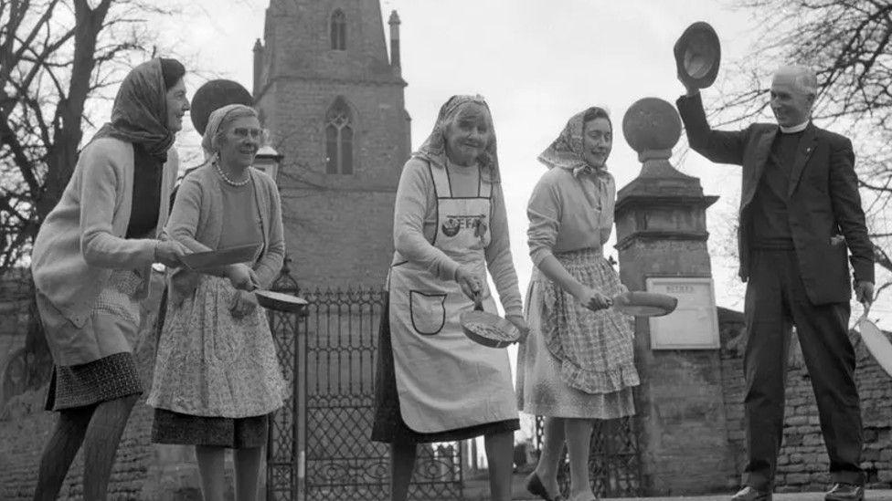 Four women dressed in skirts, aprons and headscarfs each carrying a frying pan with a pancake inside. On the right is the Reverend Canon Ronald Collins who is waving a hat in the air signalling for the women to start running.