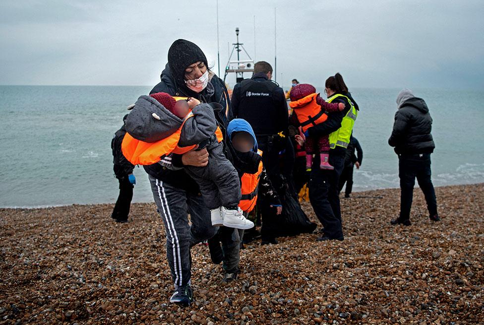 A migrant carries her children (with faces blurred) after being helped ashore from a Royal National Lifeboat Institution lifeboat