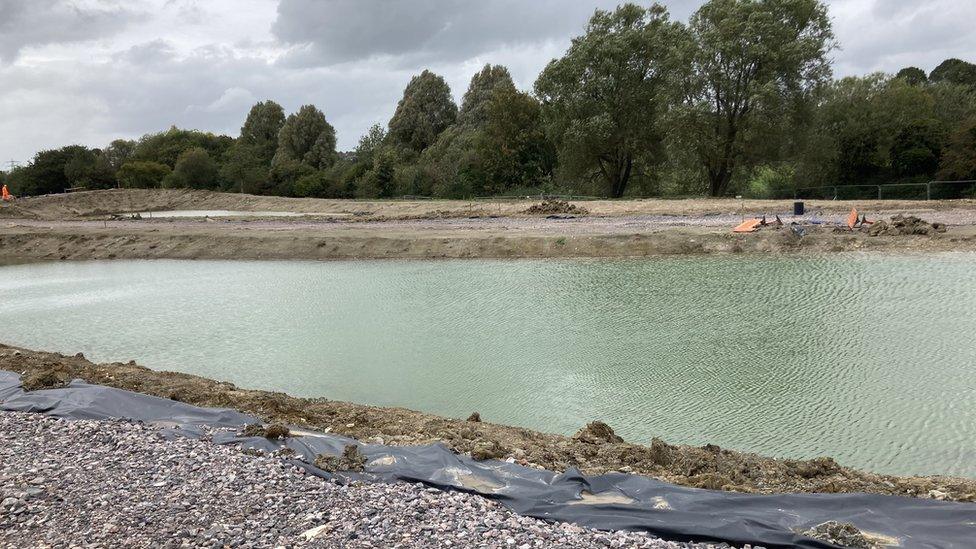 A construction site with a large section of water in the middle. Surrounded by trees.