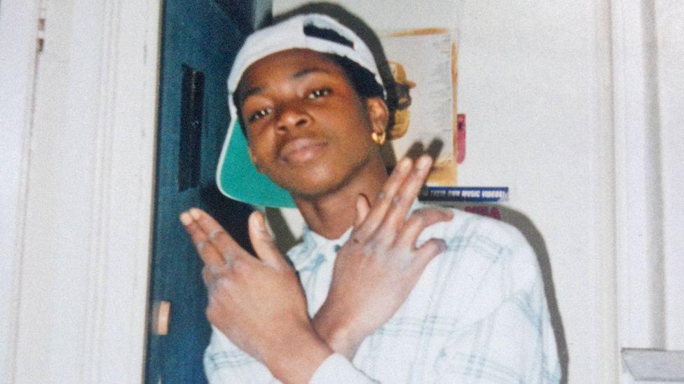 Mark Wilberforce smiles at the camera as a teenager in London wearing a white cap backwards, a gold earring and a white checked shirt. His hands are crossed and held up with the last two fingers on each bent.