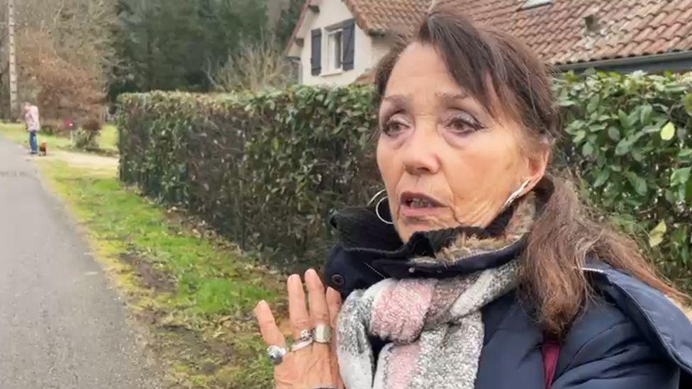 A woman with brunette hair, wearing a coat and scarf, is looking away from the camera. She is stood outside and appears to be mid-conversation