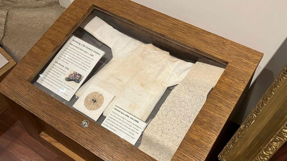 A wooden display box and under its glass is a formerly white baby's gown or shirt, lying on its back with its arms outspread. On the far right is a length of intricate Flemish lace.