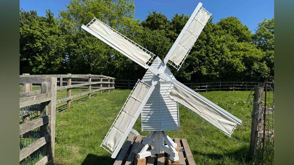 A white replica of Bourn Windmill. It has white sails in an X-shape attached to its peaked roof. It has a white slatted body and is resting on a white X-shaped base on a pallet. Behind the model is grass and trees. 
