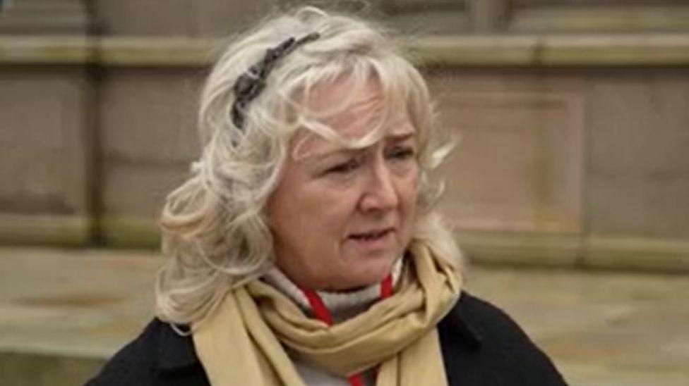 Deputy Karen Wilson is wearing a black jacket and a beige scarf. She has blonde hair and is stood outside a stone building