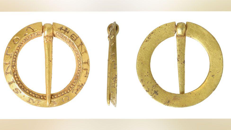 Three views of a small, gold medieval brooch. The front is made from a ring with a gold pin crossing over the middle to touch the other side of the ring. The outer ring has words etched into it and the inner ring has a decoration of tiny gold dots. The back side is plane and the third view is of the brooch's side