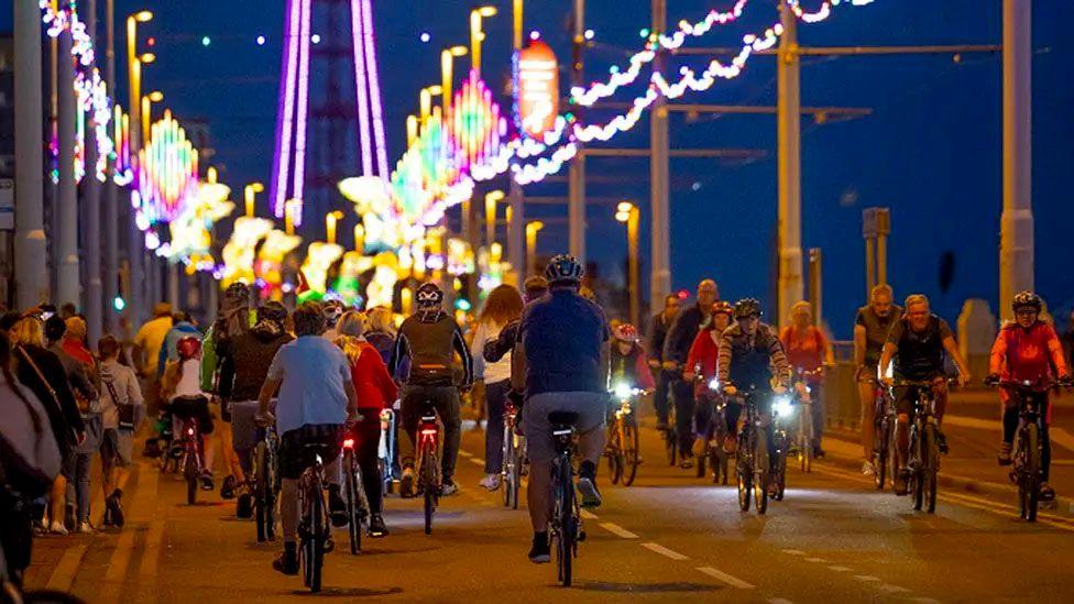 Blackpool prom at dusk with dozens of cyclists seen at a distance riding under Blackpool Illuminations