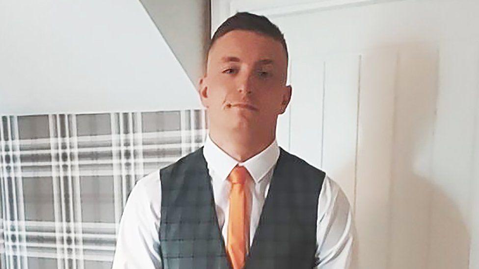 Michael Jenkins, man wearing white shirt, a waistcoat, and an orange tie, looking at camera, with an interior wall behind him