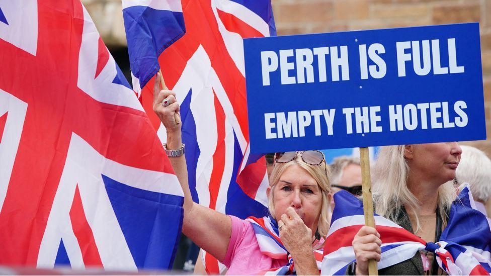 People demonstrating at an Abolish Asylum System protest outside the Radisson Hotel in Perth. Two blonde women are wrapped in Union flags and one holds a sign which reads "Perth is full empty the hotels"