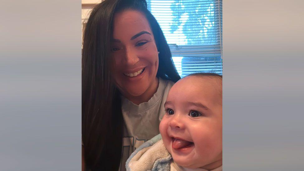 Sasha Field with long black hair, who is smiling as she is holding a baby who is also smiling with his tongue sticking out.