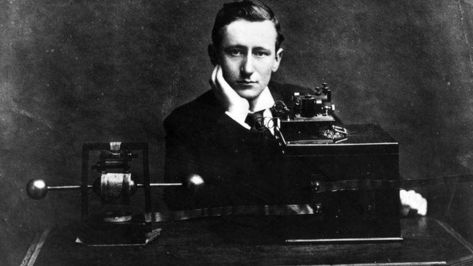A black and white image of Guglielmo Marconi wearing a suit and leaning on his right hand. In front of him are large pieces of wireless equipment on a desk.