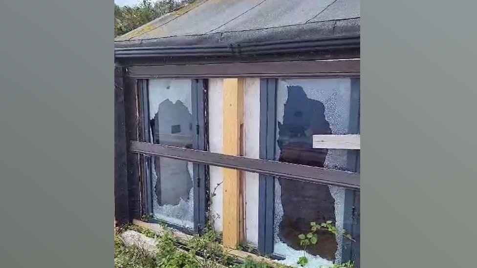 A close-up of black framed two-pane window with both panes smashed, showing through into a room beyond