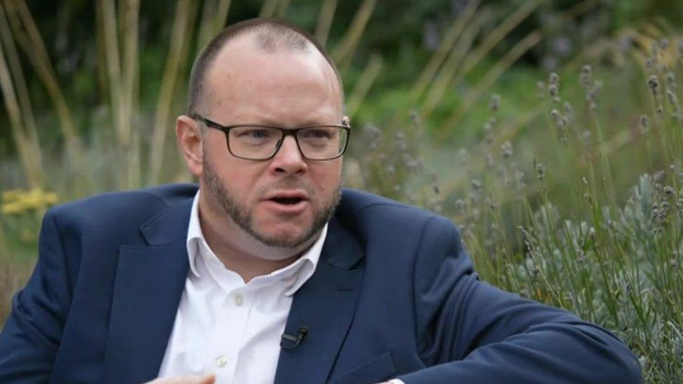 A middle-aged man with dark glasses, a short chinstrap beard and short hair wears a blue suit jacket, white shirt and looks at the camera from a park bench.