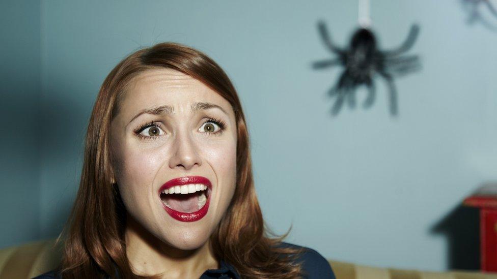 staged image of woman in bright red lipstick looking scared as a large black spider descends from the ceiling. The room is blue and she has brown eyes and hair