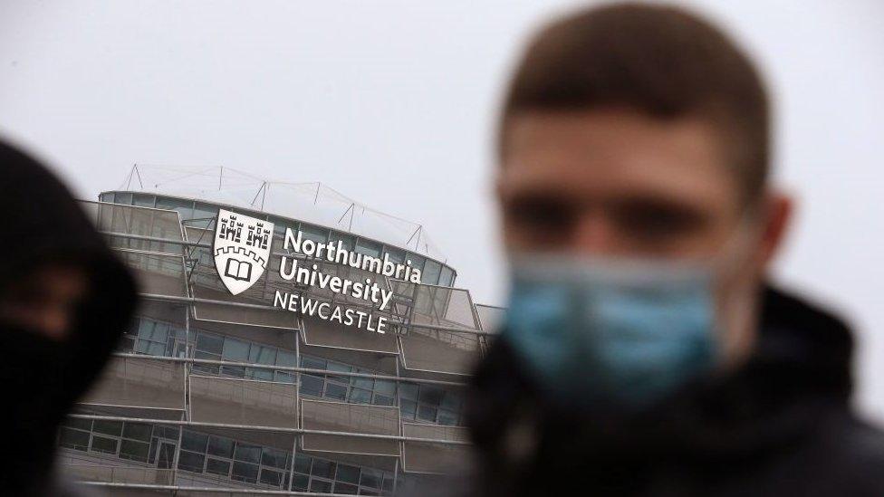 A student wears a mask in the foreground with a Northumbria University building behind him