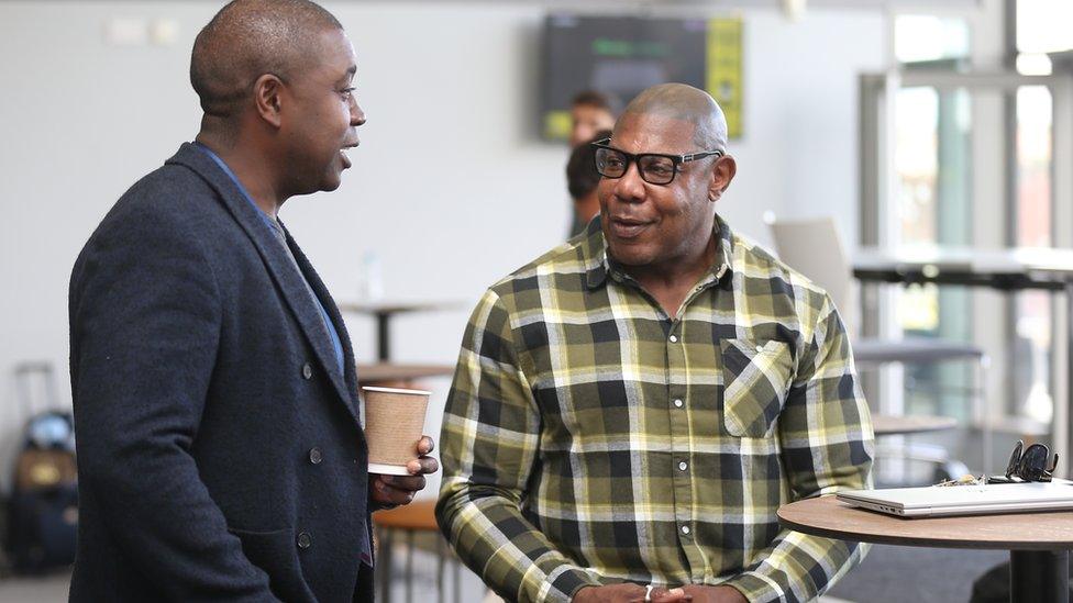 Two men talking to each other, one of them wearing a green checked shirt