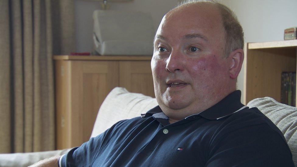 John Clarke sits at home in a blue shirt