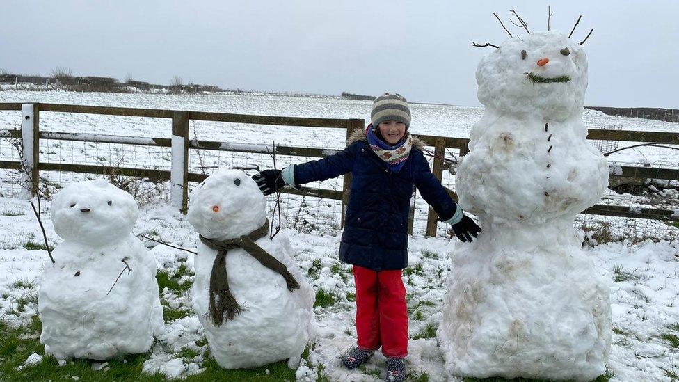 A child stands in the middle of a row of three snowmen with their arms held out to the side