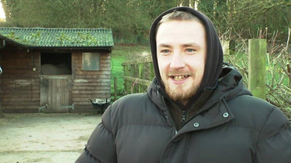 Patrick. He is wearing a black coat. He is stood near the horse stables, smiling.