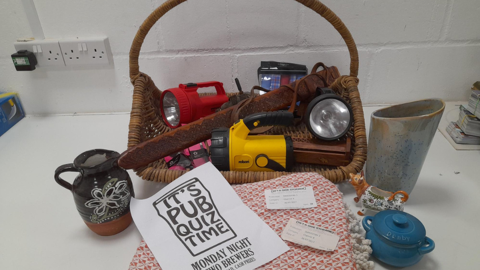 The props inside a basket on a white counter top. They include large yellow,red and black torches. A sword sheath and crockery.