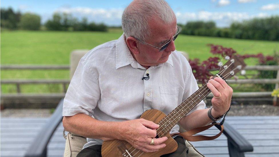 A man with white hair in a white shirt holding a ukulele