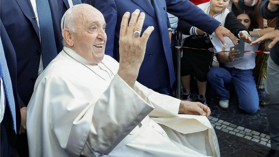 Pope Francis in a wheelchair smiling and waving to people as he leaves hospital in Rome