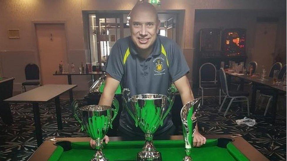 Scott Lloyd leaning against a pool table. Trophies are shown.