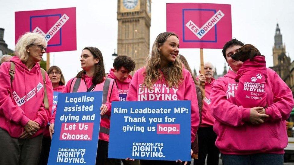 A group of campaigners in favour of legalising assisted dying are dressed in pink hoodies which say "campaign for dignity in dying", and hold placards which read "yes to dignity" at a rally outside the Houses of Parliament.