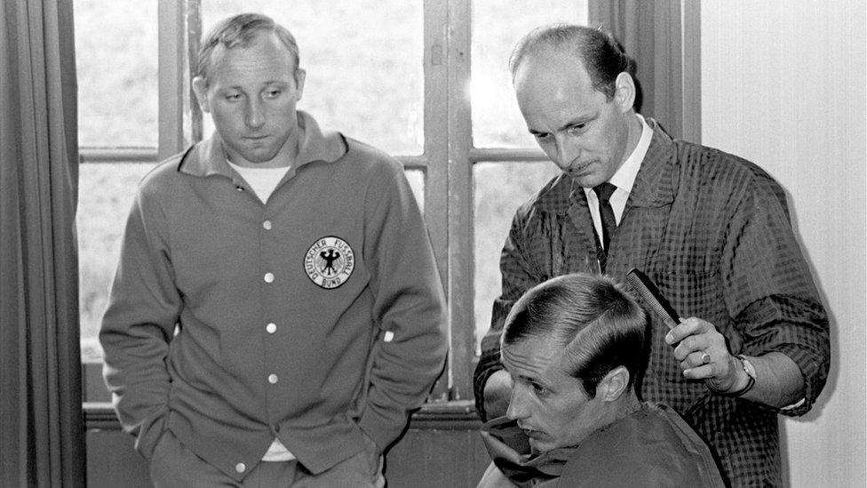 West Germany's Uwe Seeler (left) looks on as teammate Heinz Hornig (right) is given a trim by a local barber at the team's hotel in Derbyshire during the 1966 World Cup (14 July 1966)