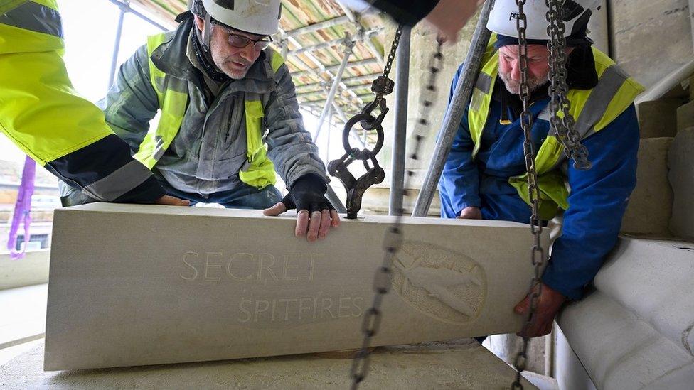 Men wearing high-vis and hard hats putting a stone with a spitfire carving in place