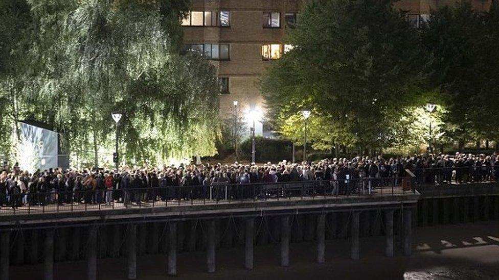 People queueing along the River Thames at night time.