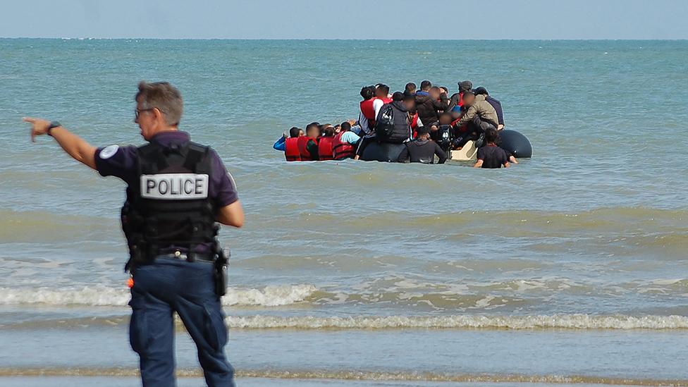 A picture of a French police officer in the foreground, gesturing to migrants on a small boat in the shallow sea in the background