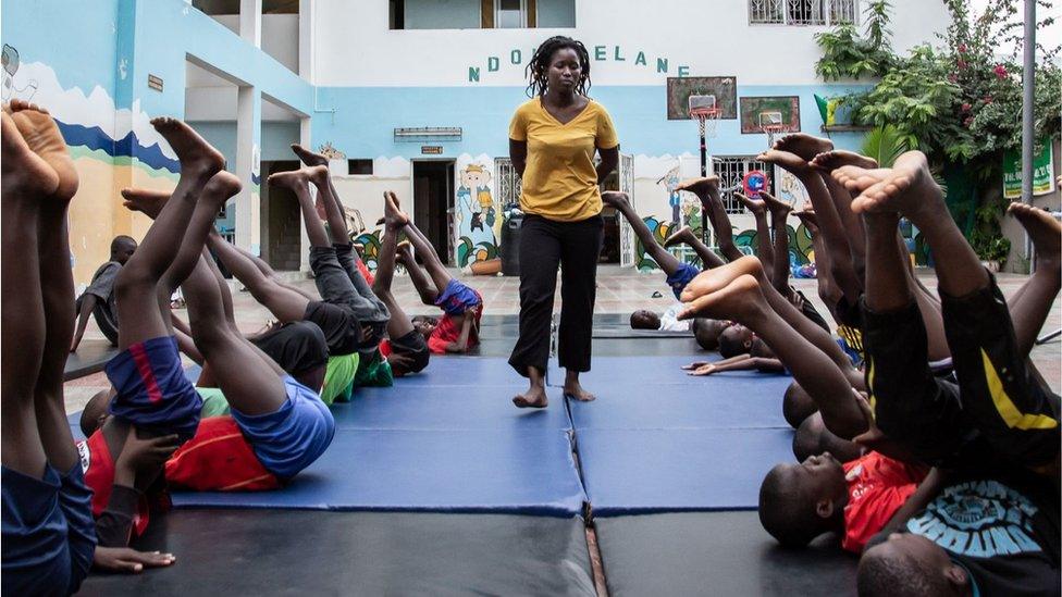 A woman walking through two lines of acrobats who appear to be practicing. They are on their backs with legs in the air.