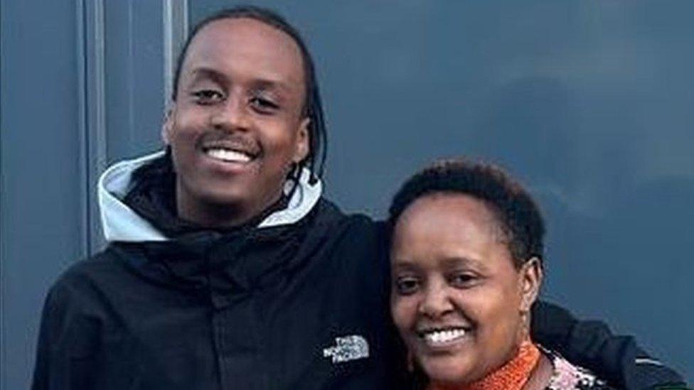 A young black man and a middle aged black woman with their arms around each other smiling