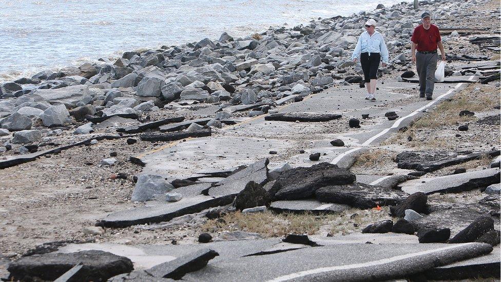 Two people walking along a road that has been almost completely destroyed, with rubble and pieces of tarmac everywhere