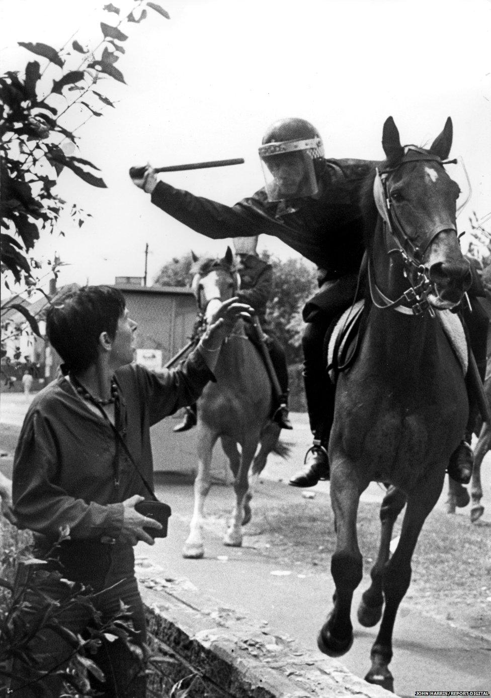 Police officer on horseback aims truncheon at woman in crowd