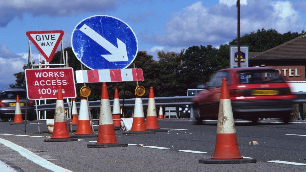 Car drives past a "works access" sign and orange cones