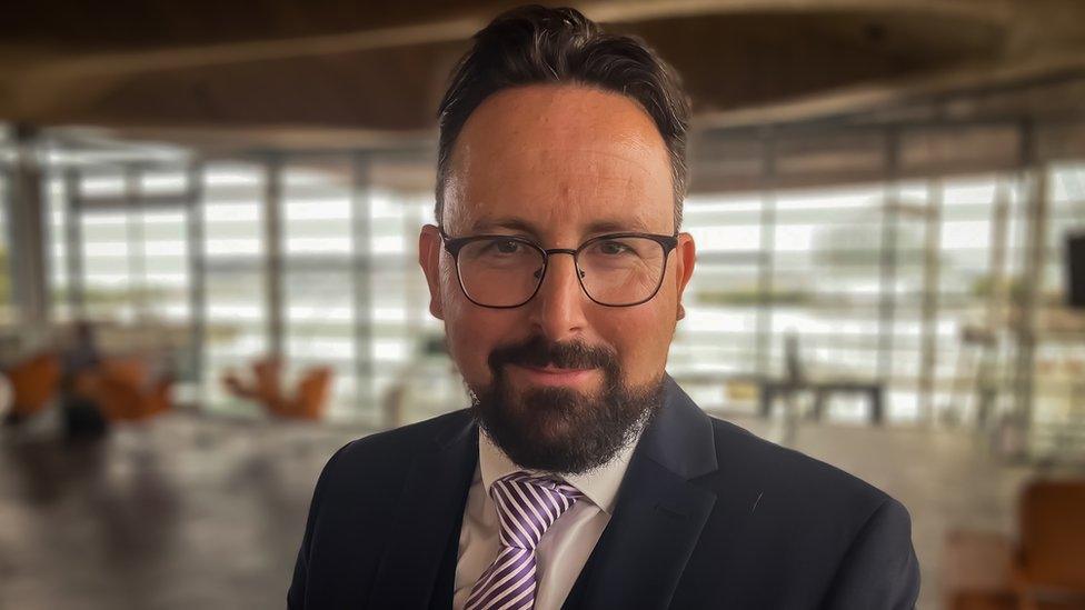 Hefin David standing in a foyer, wearing a suit and tie