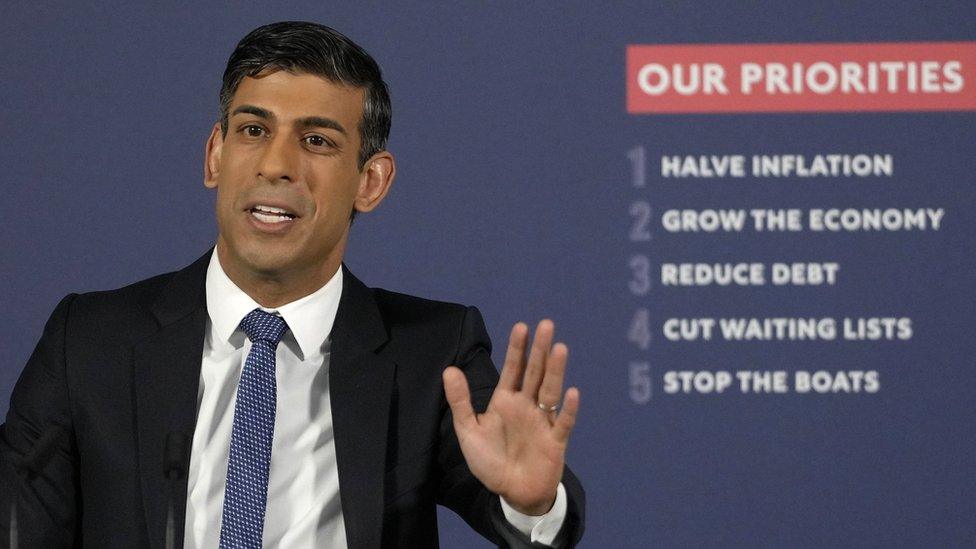 Rishi Sunak in front of a board with his government's five priorities on it. He has his left hand raised