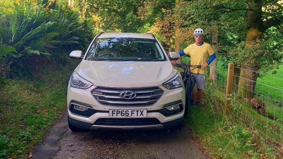 Narrow road with a car filling the entire width. A cyclist is squashed at the side.