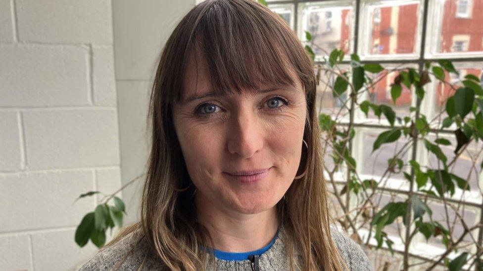 Image of Sophie Wilsdon. She has dark blonde hair and is wearing a grey jumper. She is looking directly at the camera and smiling. A large window and a plant can be seen in the background behind her.