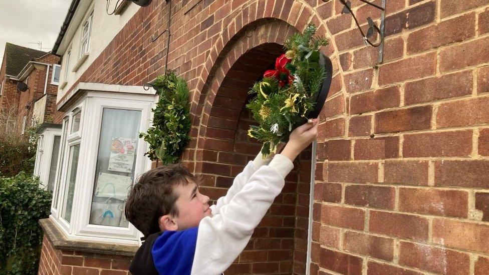 Boy dressed in blue and white reaches up to hang a holly wreath beside a front door