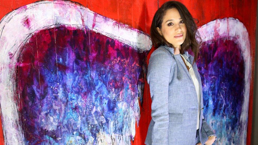 Meghan Markle poses with angels wings painted in bright colours on a wall behind her