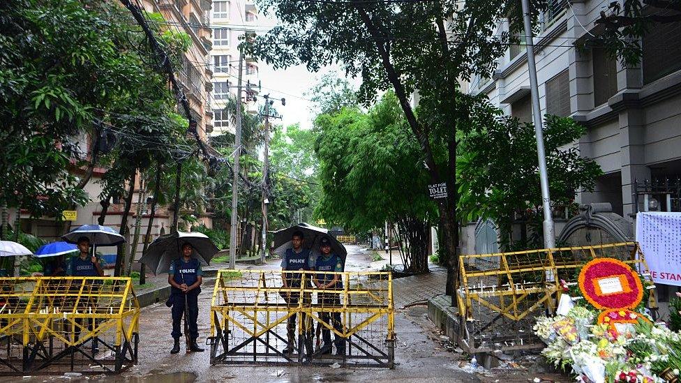 Bangladeshi policemen stand under umbrellas at a checkpoint in Dhaka on July 5, 2016, in a street leading to the entrance of a restaurant which was the site of a bloody siege that ended in the death of seventeen foreigners and five Bangladeshis.