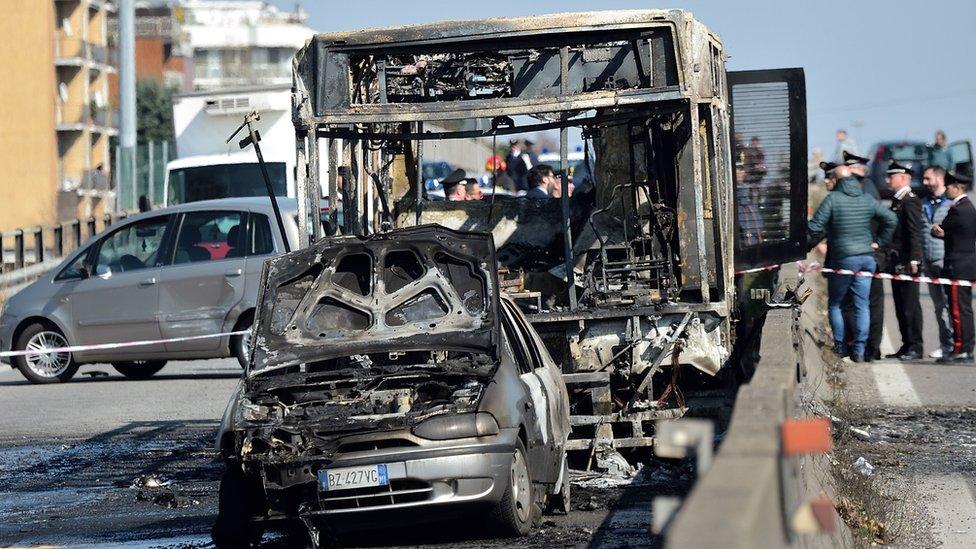 A burned-out bus is seen on the road, surrounded by uniformed police, in the aftermath of the fire.